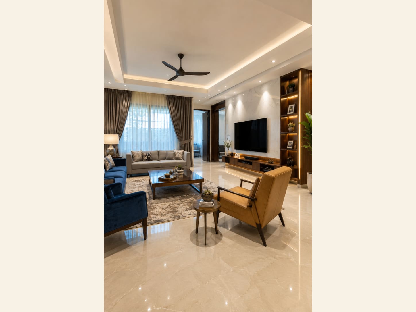 A light-filled living room with a cream sofa, navy velvet armchairs, a tan leather chair and a marble TV wall with warm-lit wooden shelving.