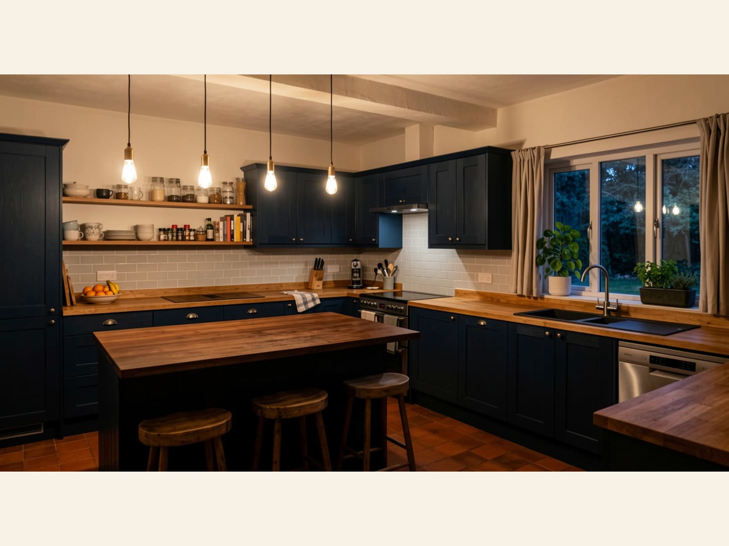 A cosy English country kitchen with deep navy shaker cabinets, warm butcher block worktops, open wood shelves, Edison bulb pendants and terracotta floor tiles.
