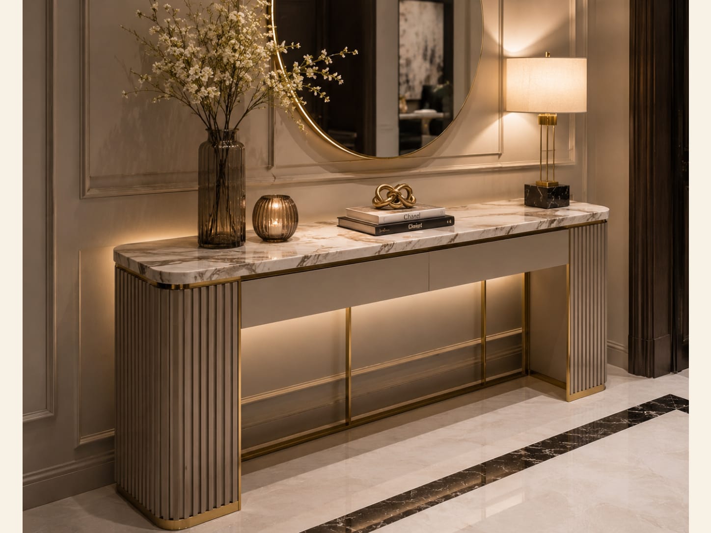 A Greige Fluted Console with a Marble Top and Round Gold Mirror A greige fluted-base console with gold trim and a white marble top, a large round gold mirror on a panelled wall, a marble lamp, a gold knot sculpture and a dark marble floor inlay.