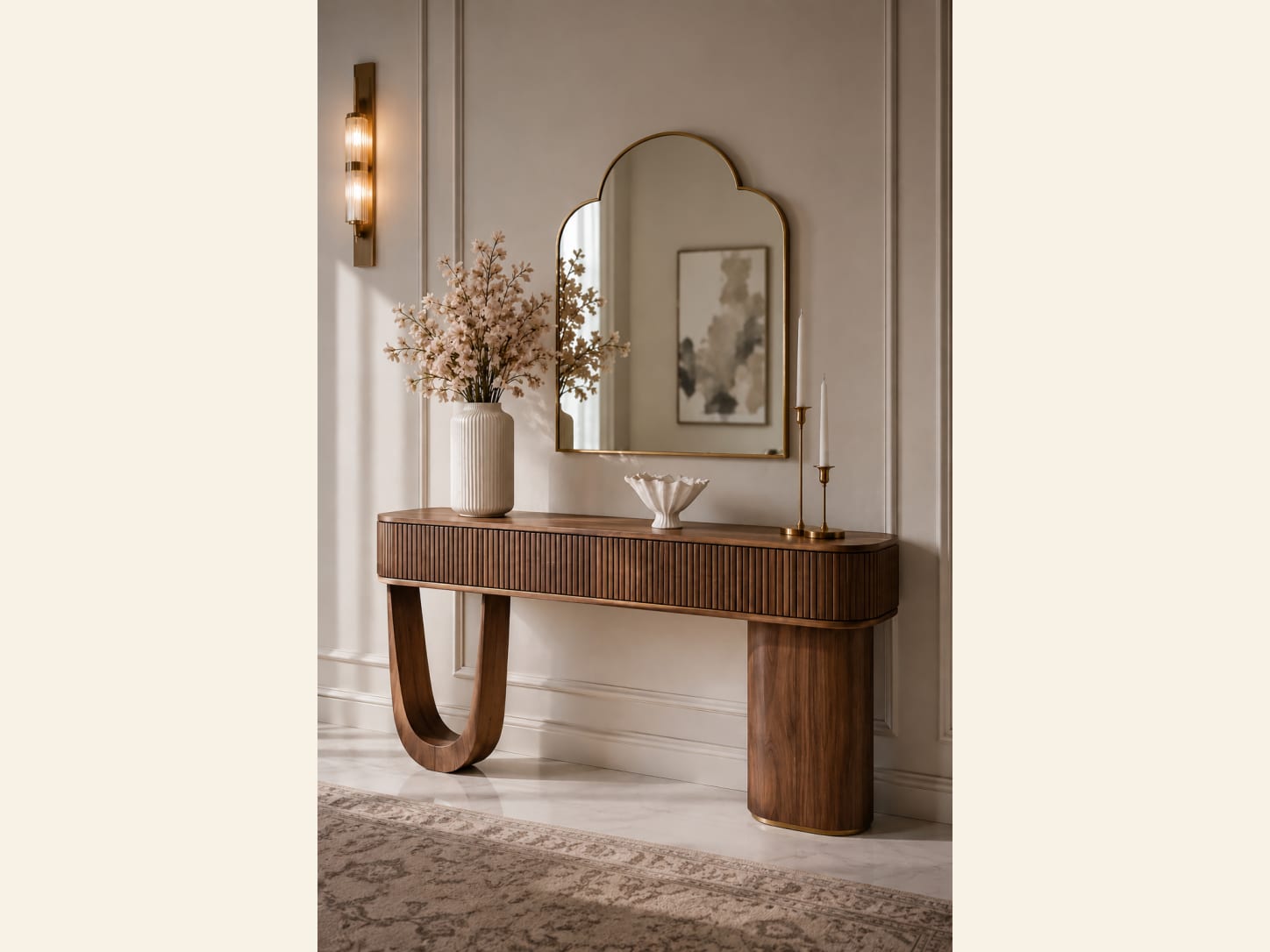 A Fluted Walnut Console with an Arched Mirror An elegant entryway with a fluted walnut console on a curved loop base, an arched brass mirror, a wall sconce, a vase of blossoms and a candle on a soft panelled wall.