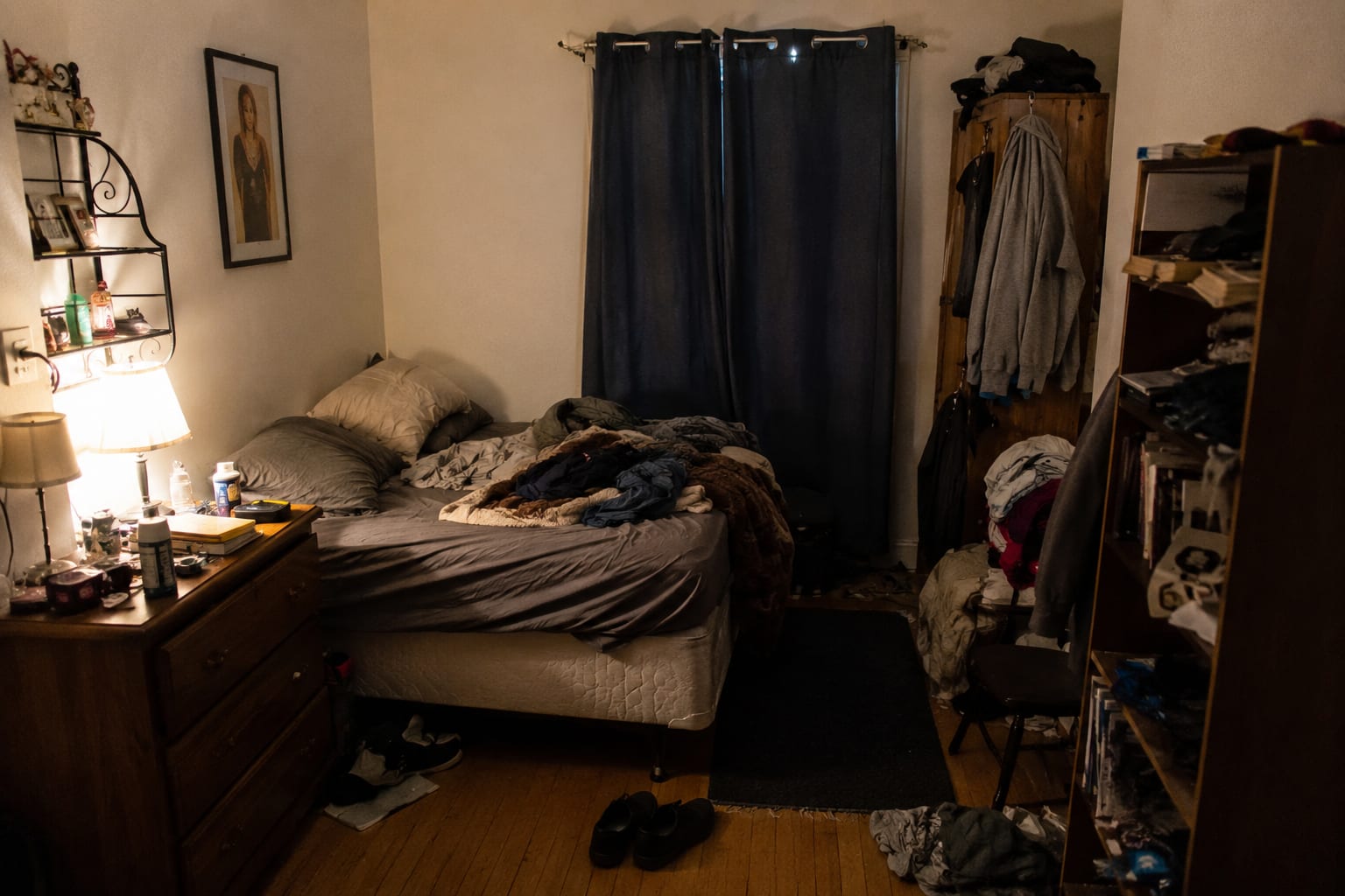 The before. Dark walls, one dim lamp, clothes with nowhere to go, and the window lost behind heavy curtains. Cluttered dark small bedroom before the makeover, messy bed, clothes piled on a chair, dim lighting