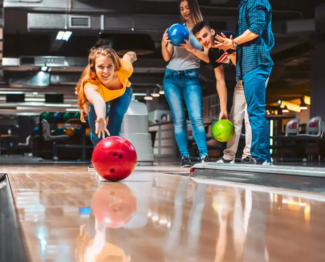 A woman bowls a red ball while friends hold blue and green balls, indoors at a bowling alley.