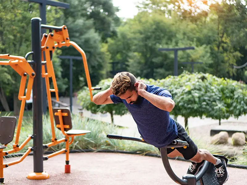A man performs an exercise on a fitness machine in a park setting surrounded by greenery.