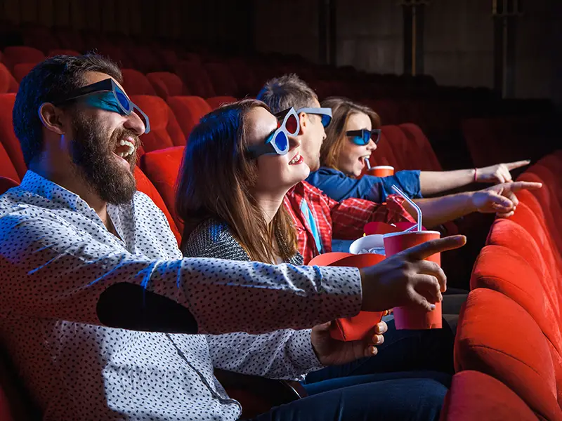 Group of four people wearing 3D glasses, laughing and pointing while seated in a theater.