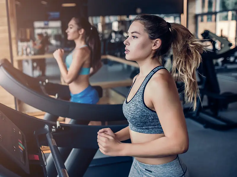 Two women running on treadmills in a modern gym with large windows.