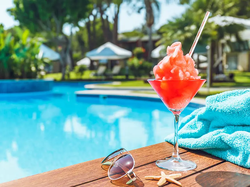 A colorful slushy drink in a glass beside sunglasses and a towel by a pool.