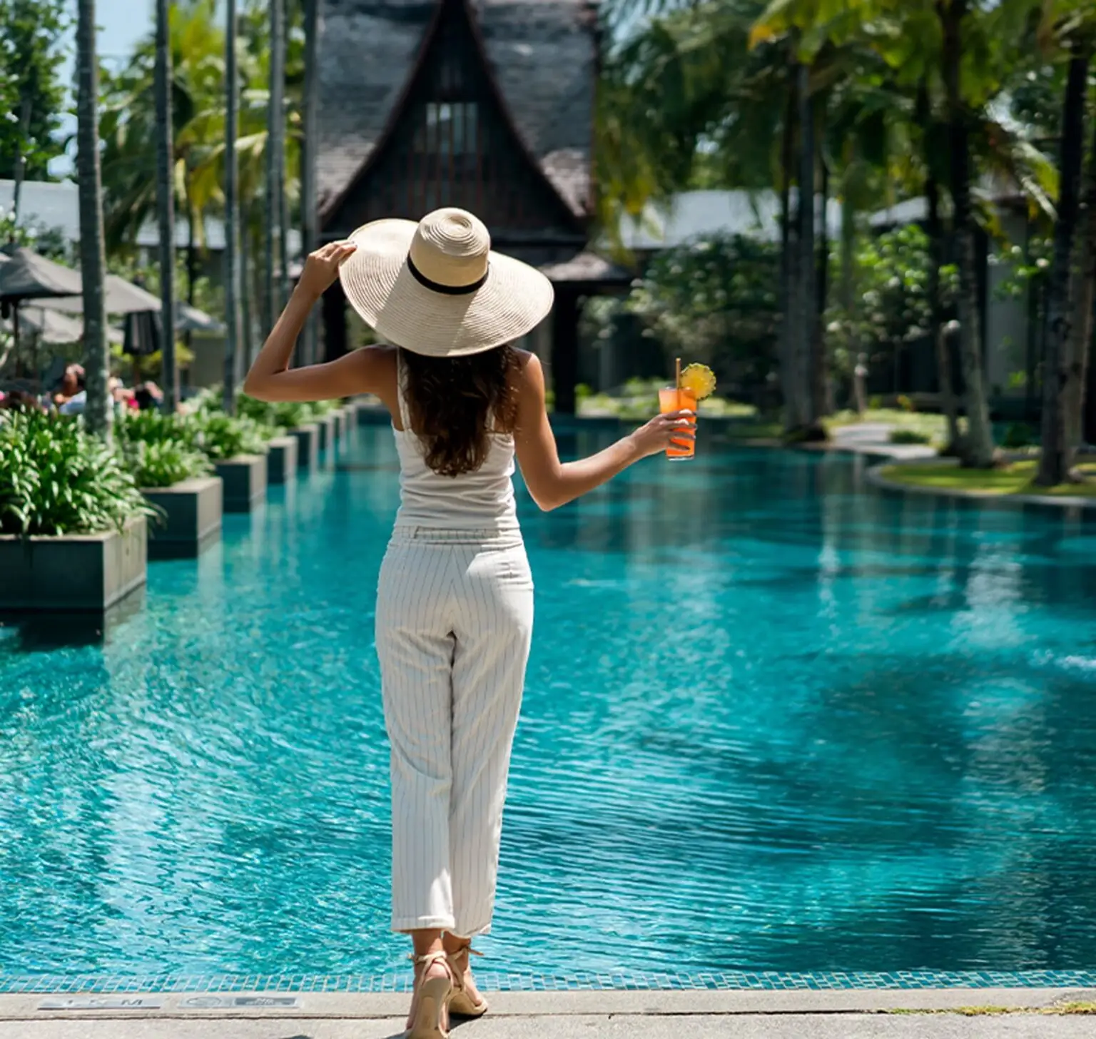 A woman in a wide-brimmed hat holds a drink while walking by a turquoise pool.