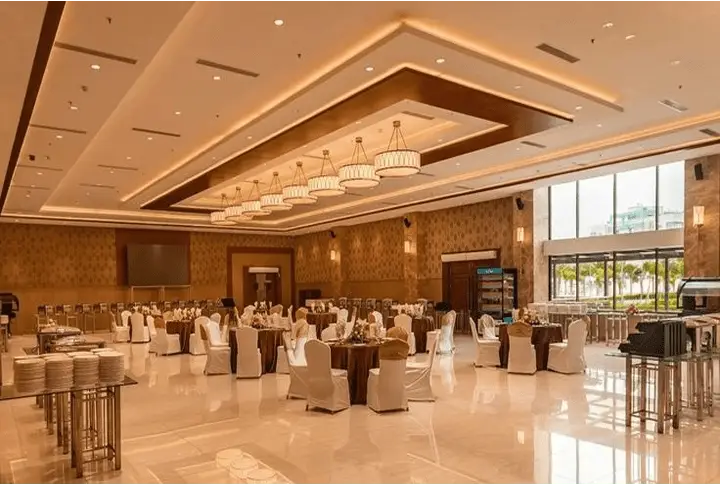 Spacious wedding hall with elegant chandeliers, round tables, and white chair covers.