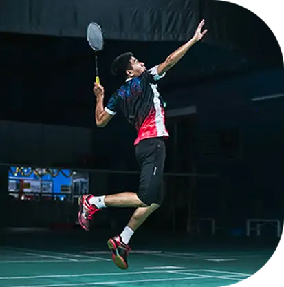 A male badminton player jumps to serve, wearing a colorful shirt and shorts.