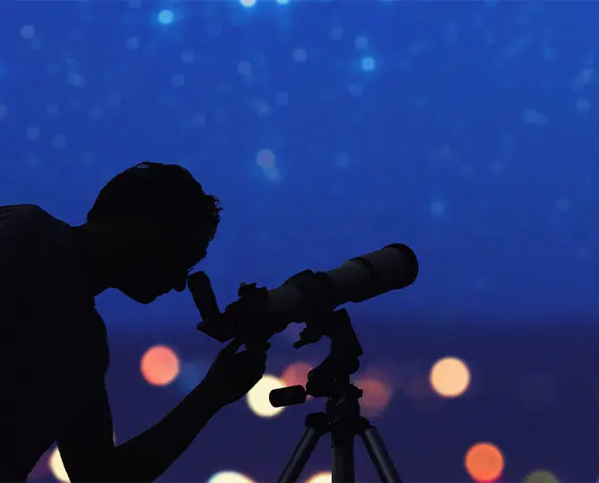 Silhouette of a person using a telescope against a starry night sky with blurred lights.