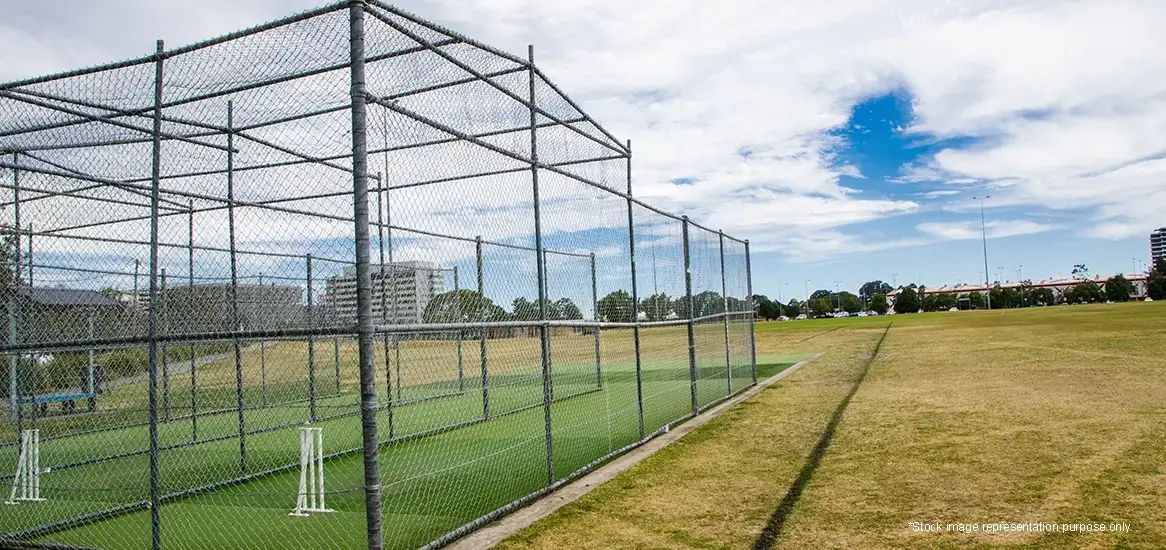 Cricket practice nets enclosed by a chain-link fence on a grassy field under a blue sky.