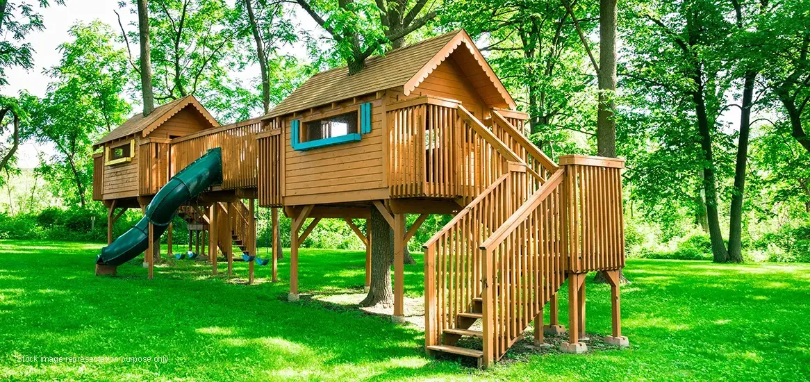 Two wooden treehouses with a slide and stairs, surrounded by lush green grass and trees.