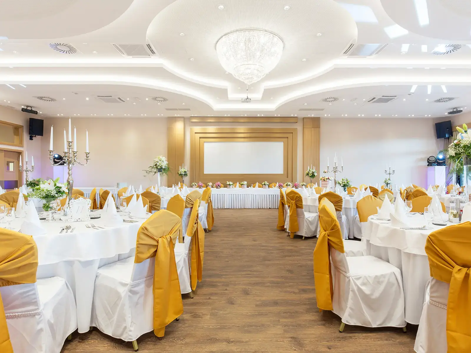 Elegant banquet hall with round tables, white tablecloths, and yellow chair sashes under a chandelier.