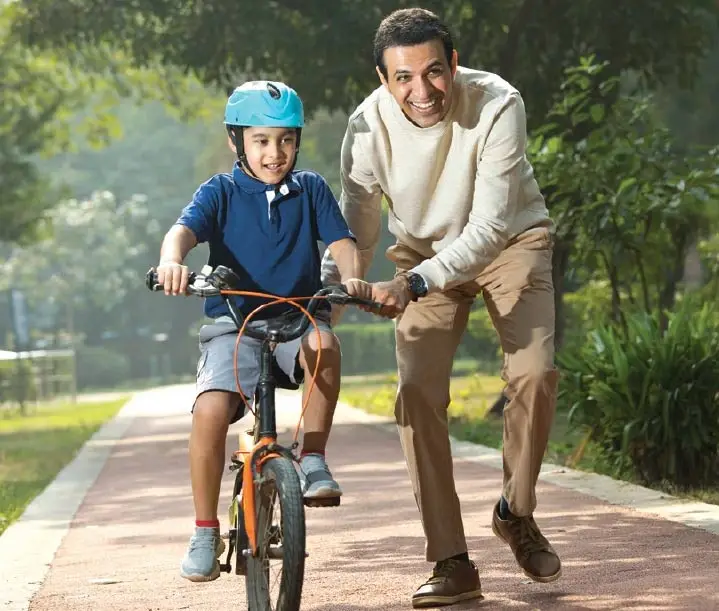 A man assists a boy riding a bicycle on a path surrounded by greenery.