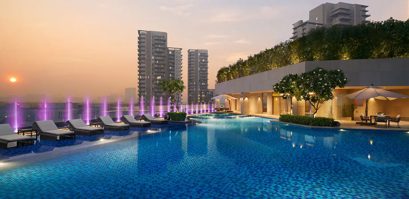 A tranquil pool area with lounge chairs, fountains, and modern buildings at sunset.