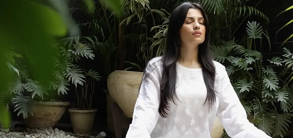 Woman in white attire meditating in a serene garden surrounded by lush green plants.