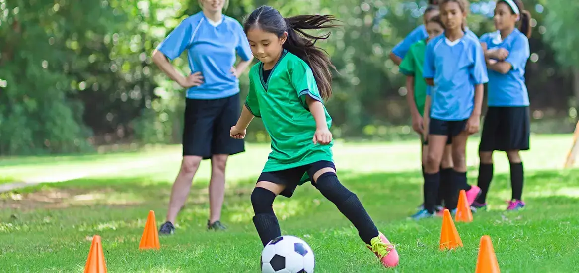 A girl in a green jersey dribbles a soccer ball past orange cones while peers watch.