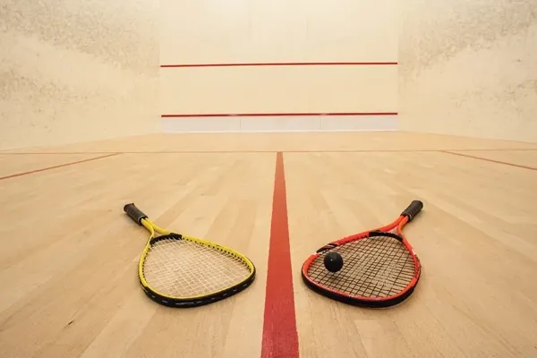 Two squash rackets, one yellow and one red, positioned on a wooden court with a black ball.