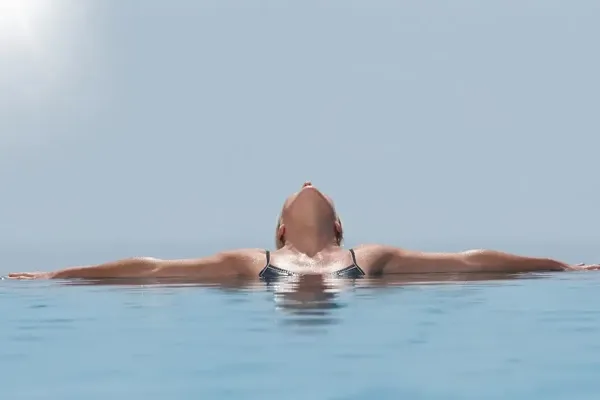 Person floating on back in a tranquil infinity pool with clear blue water.