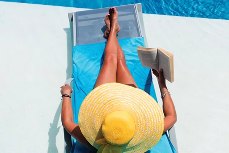 A person lounging on a blue chair by the pool, wearing a yellow sun hat and reading a