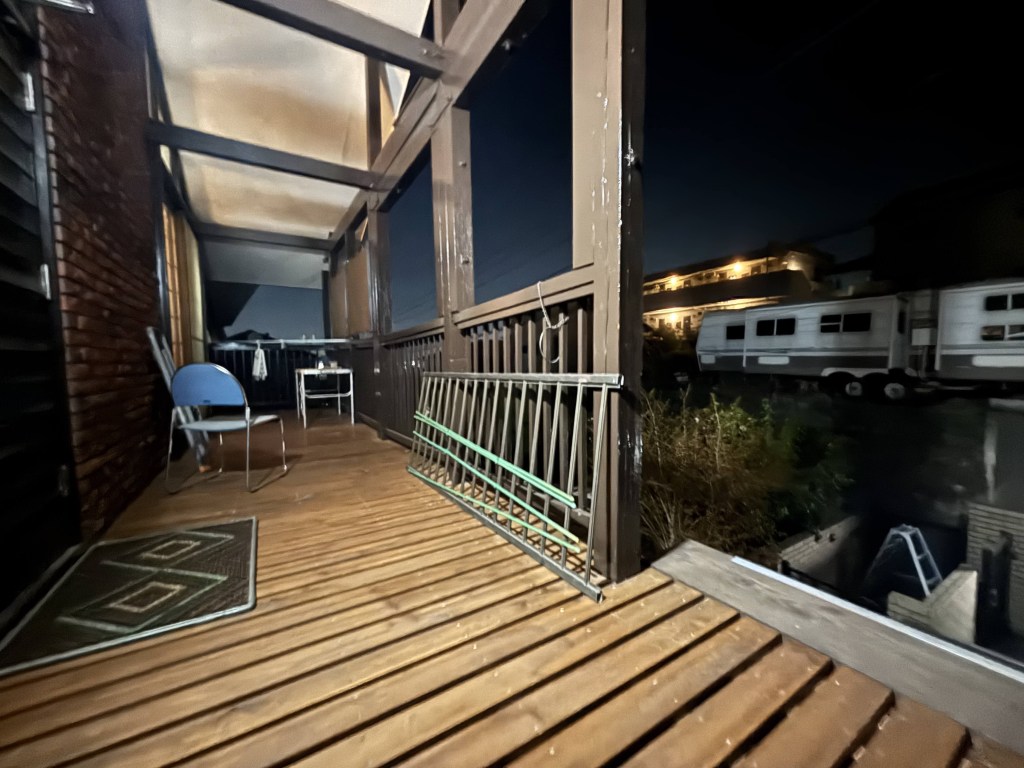 A dimly lit balcony at night featuring a wooden deck, a blue chair, and a railing with a gate. In the background, a glimpse of parked trailers and plant life can be seen.