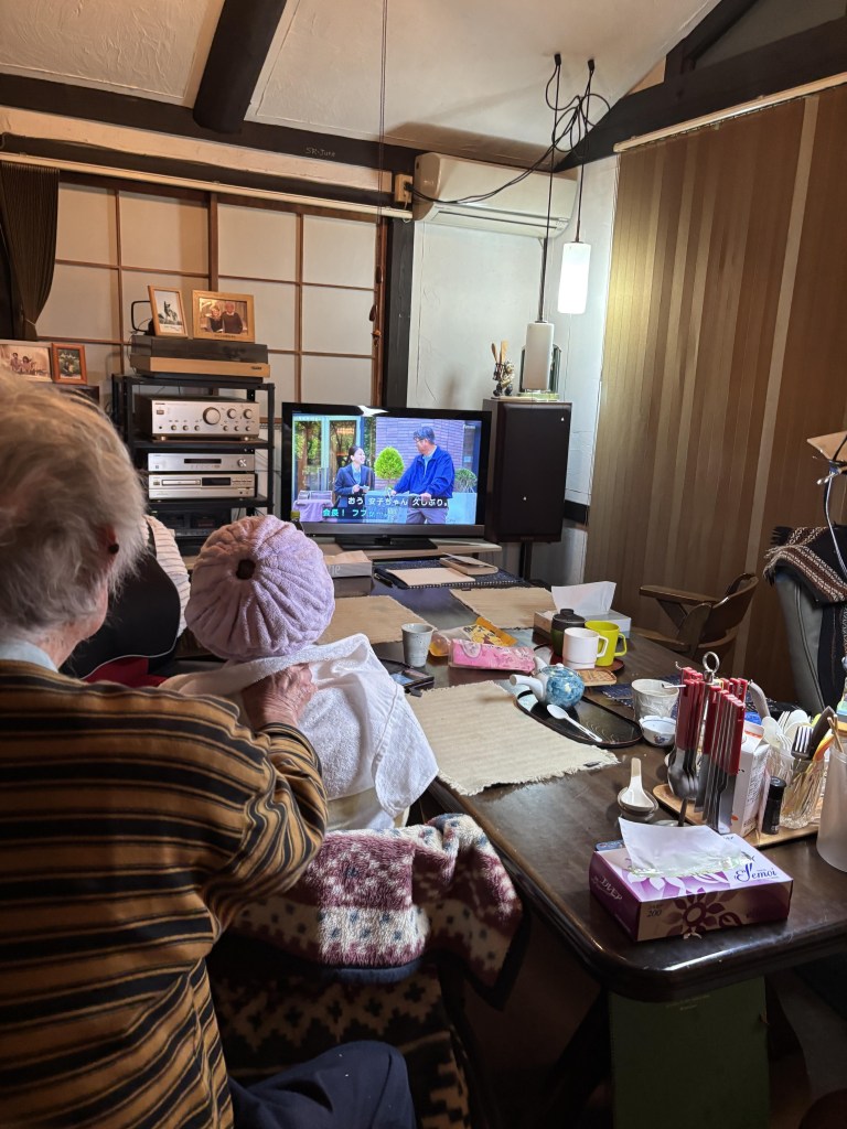 An elderly person sitting at a table watching a television show in a cozy living room, with various items on the table and a warm atmosphere.