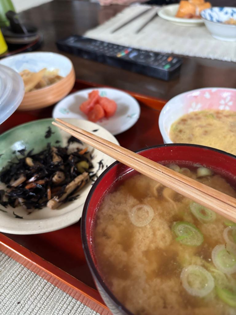 A traditional Japanese meal featuring miso soup with green onions, accompanied by various sides including sautéed vegetables and pickled items, served on a wooden tray.
