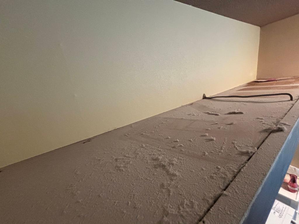 A view of a dusty, unfinished shelf with a smooth, light-coloured wall above it.