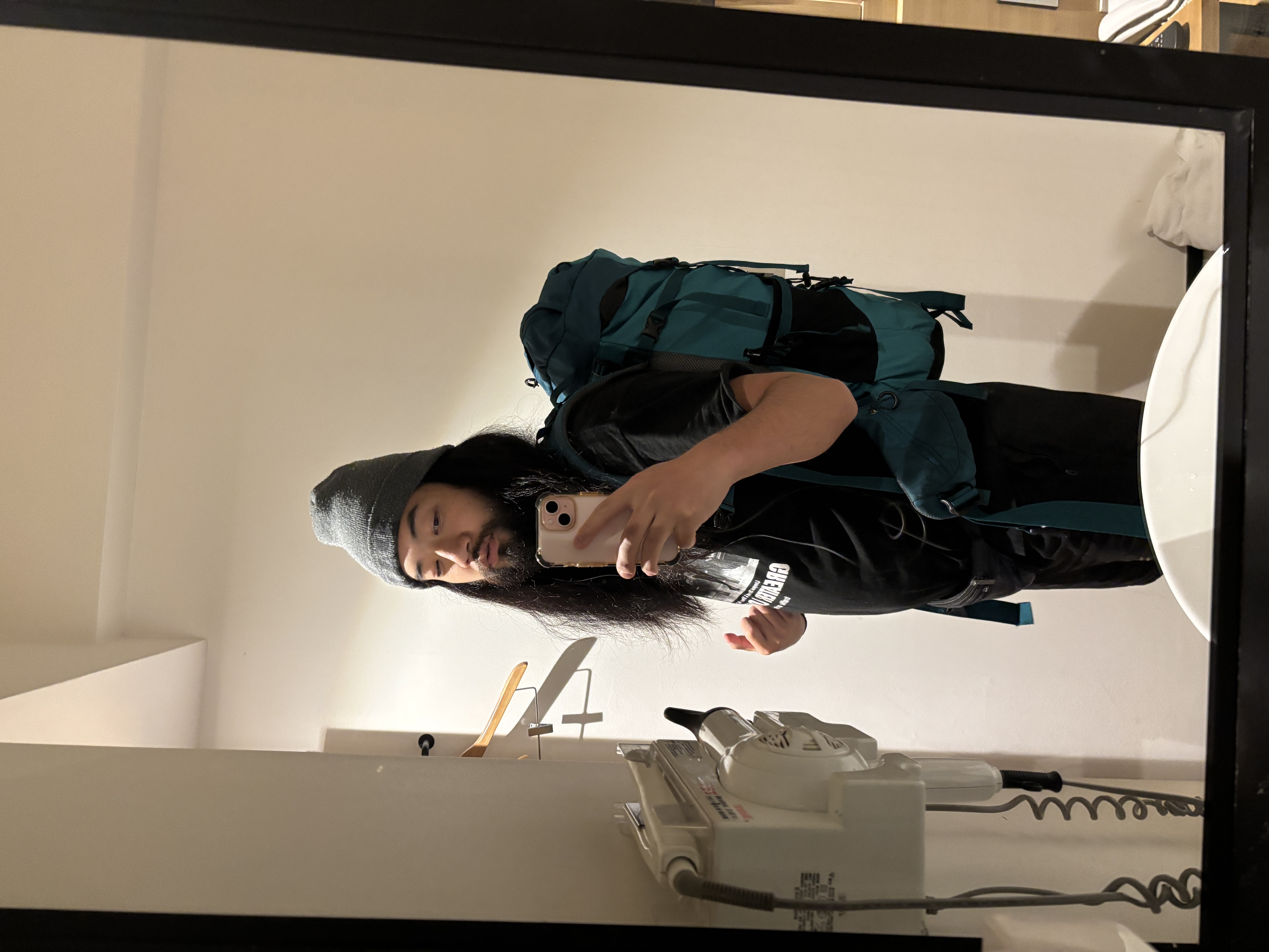 A person with long hair wearing a beanie and a black t-shirt, carrying a large teal backpack, posed in front of a mirror reflecting a tidy room.