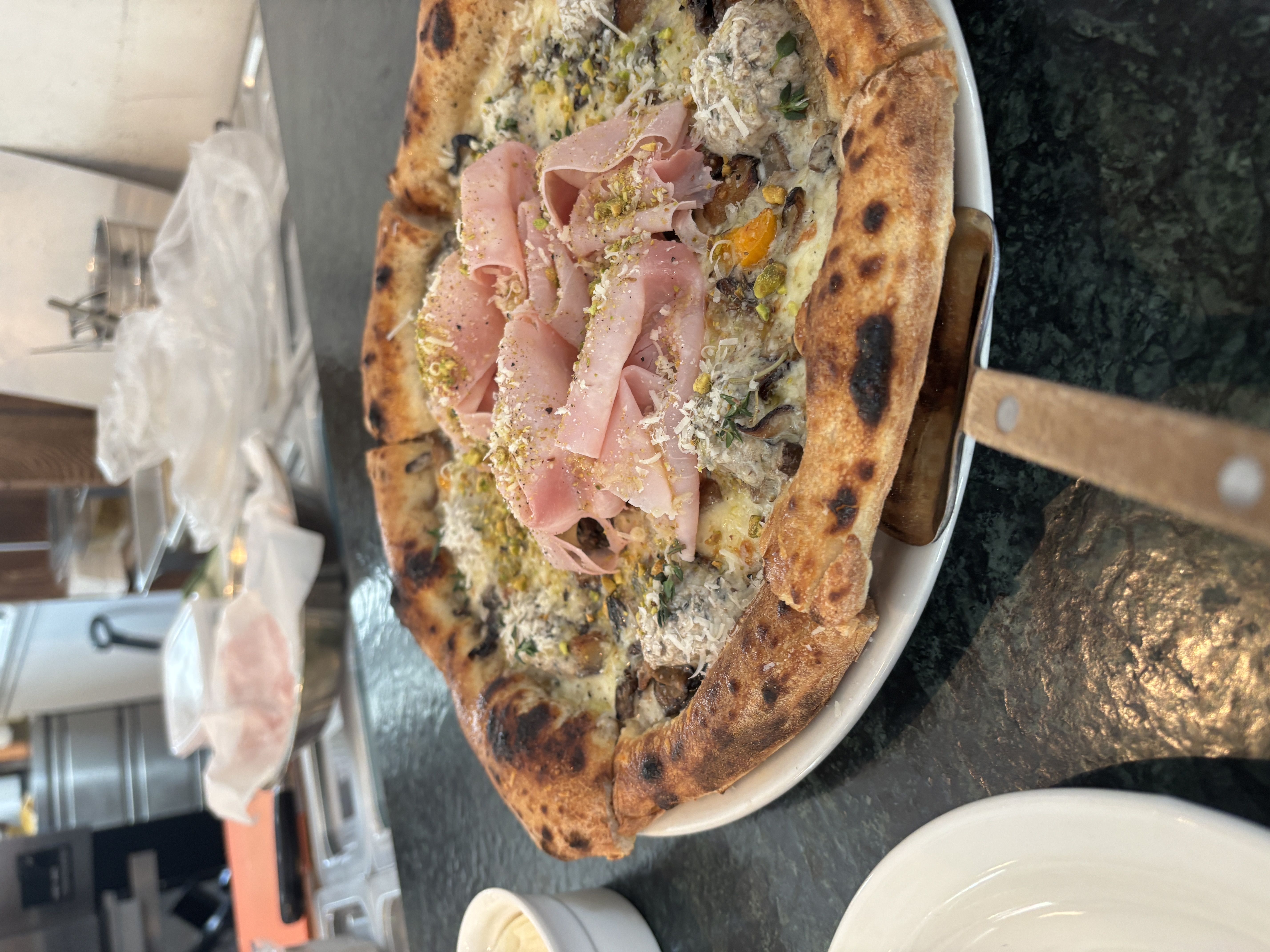 A freshly baked pizza topped with slices of ham, herbs, and cheese, served on a wooden peel with a dark stone countertop in the background.