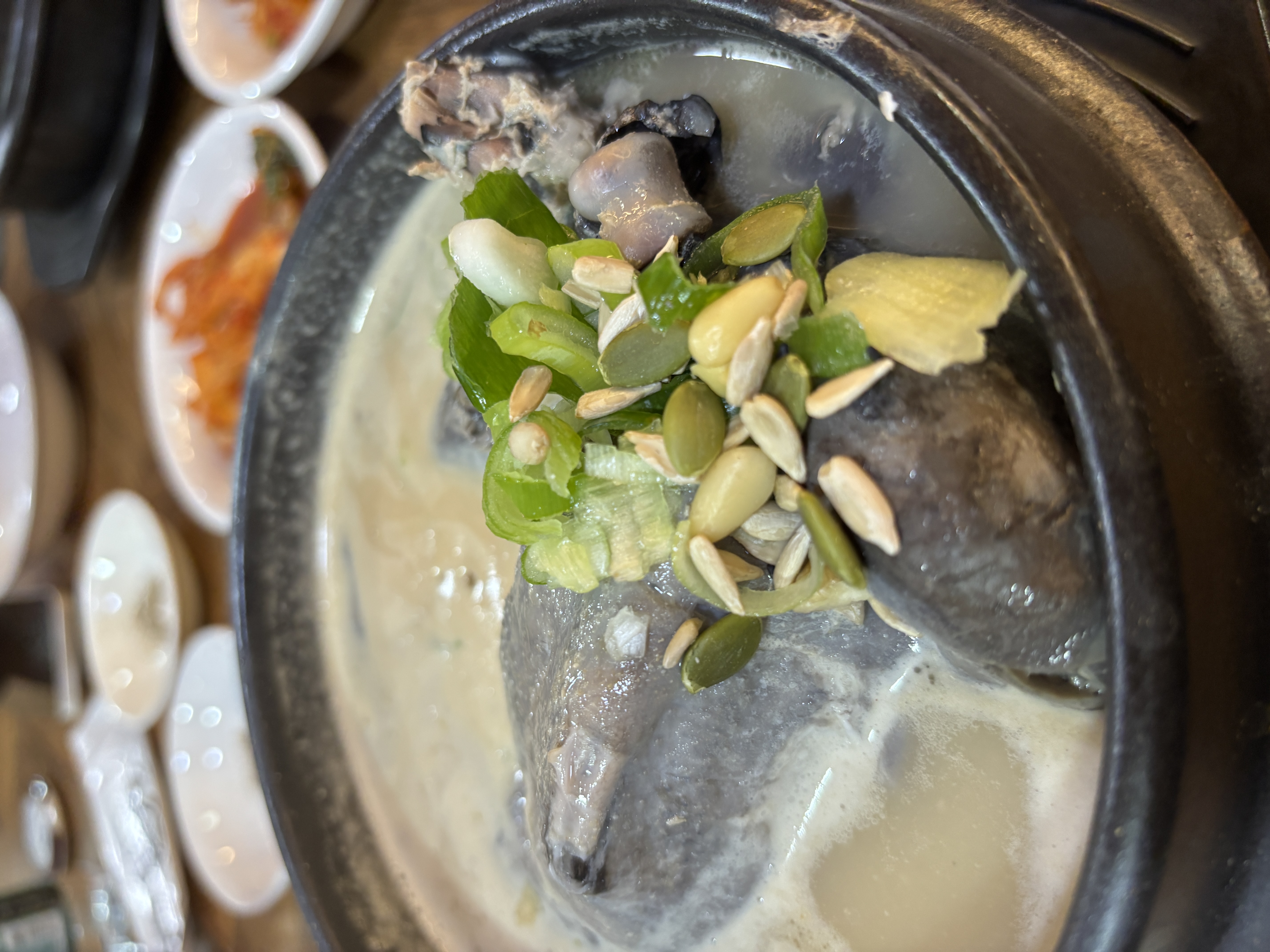 A close-up of a traditional Korean dish, showcasing a bowl of creamy soup with a whole chicken, garnished with green onions and seeds, surrounded by small side dishes.