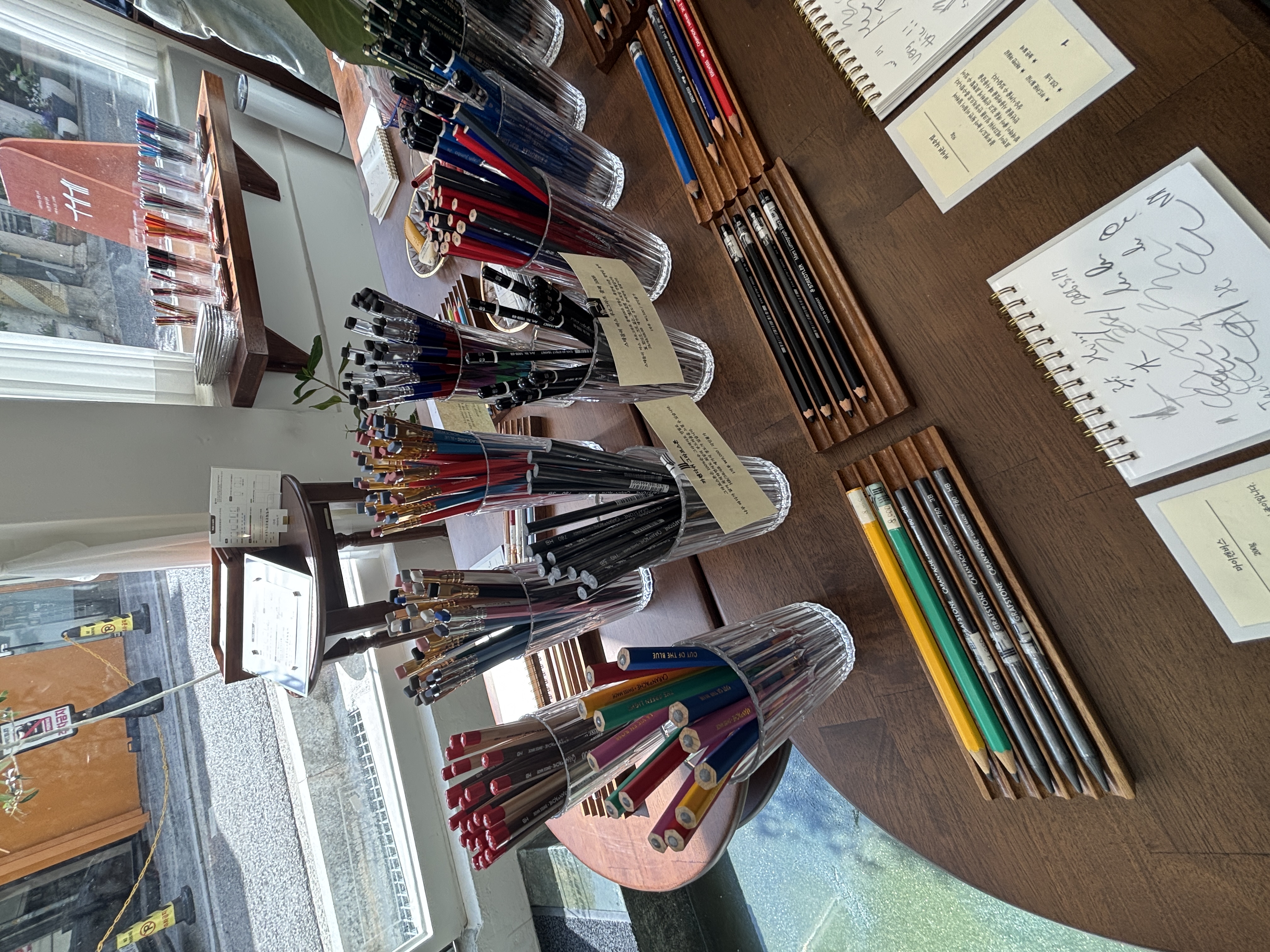 A display of various pens and pencils in clear containers on a wooden table, with notebooks and handwritten notes nearby, set in a well-lit space with large windows.