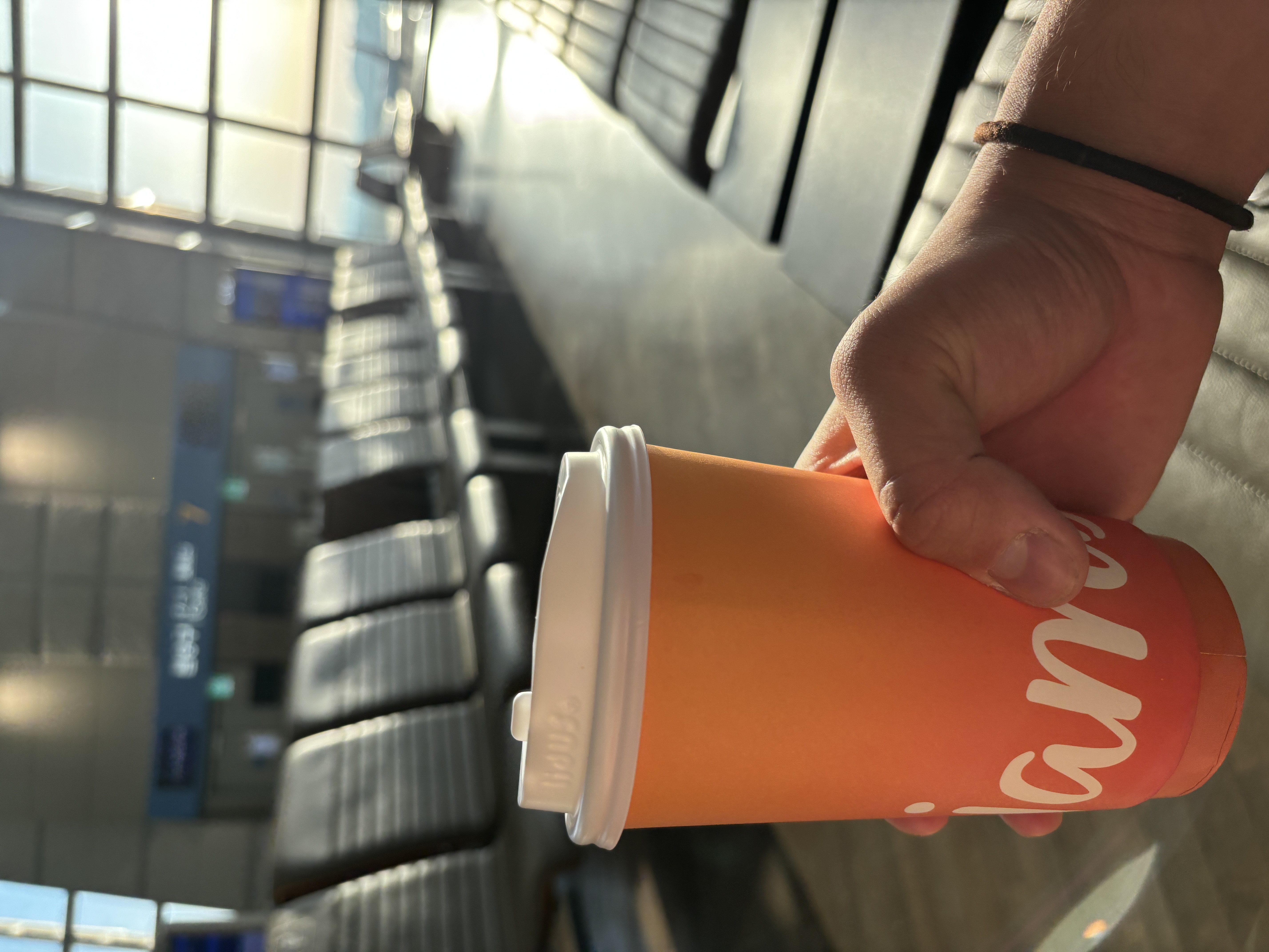 A hand holding a takeaway coffee cup with an orange gradient, in an airport terminal with empty seating and sunlight streaming through large windows.