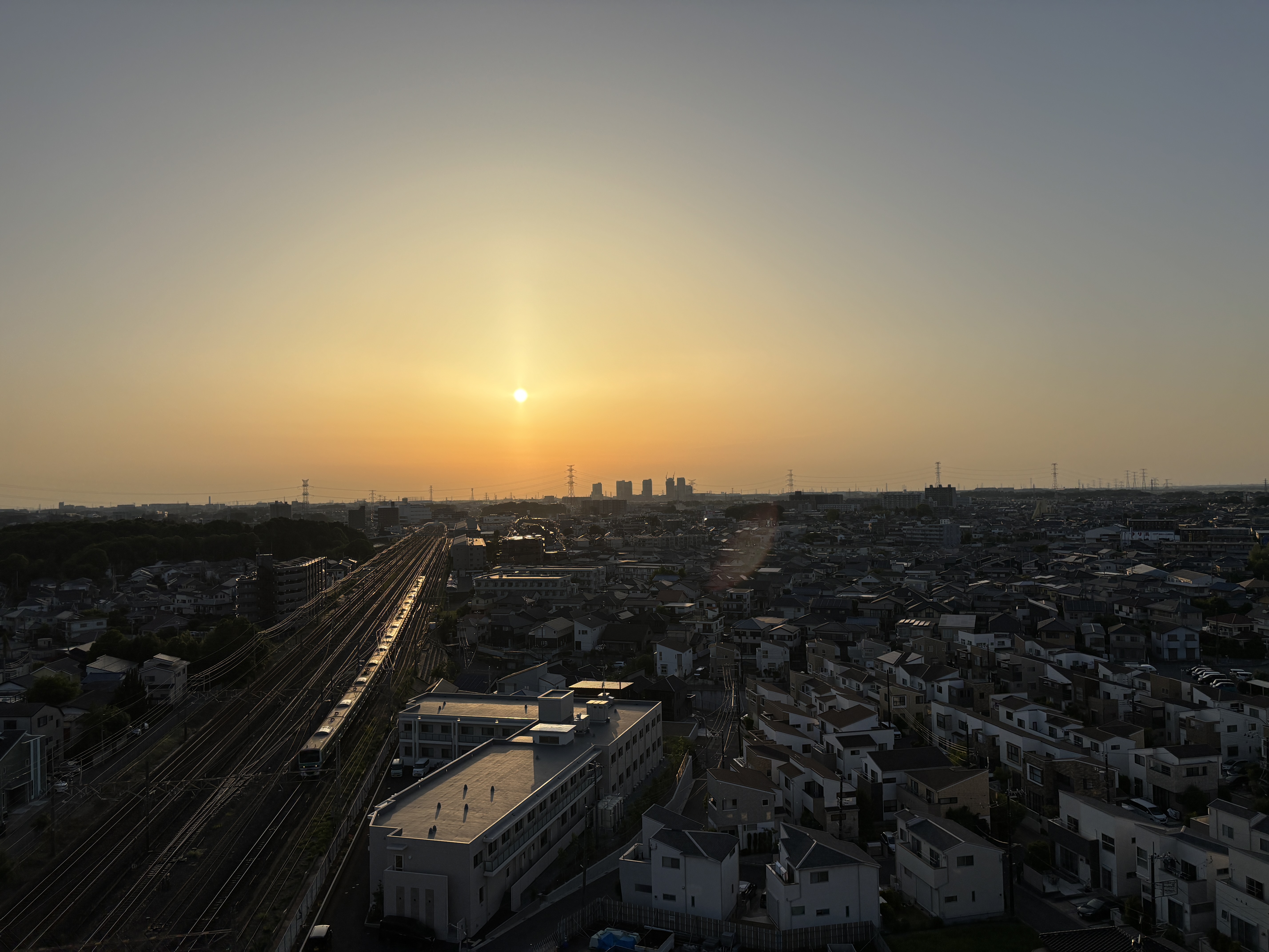 View of a cityscape at sunset, with sun low on the horizon, railway tracks winding through, and residential buildings scattered throughout.