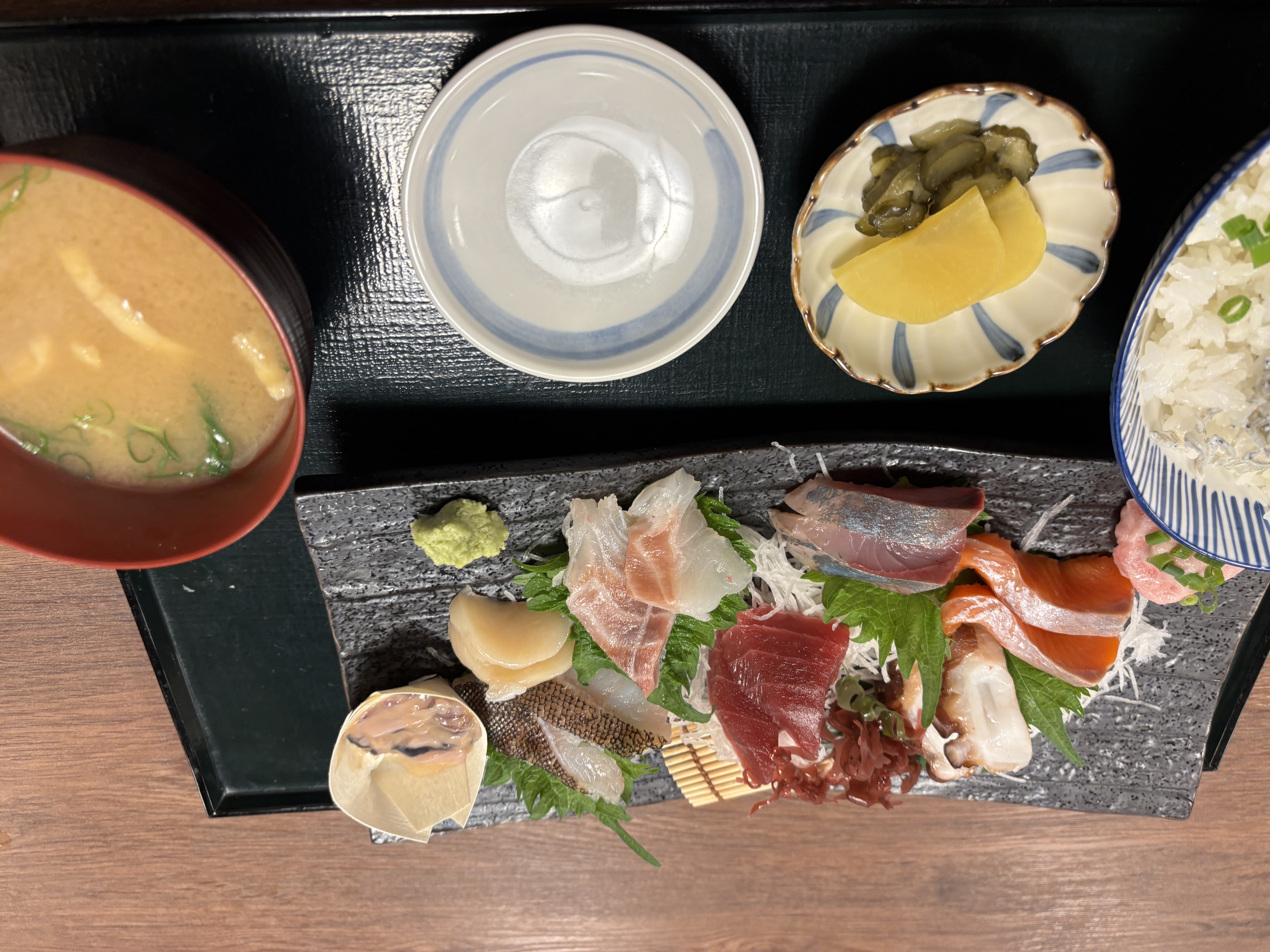 A beautifully arranged sashimi platter featuring various types of raw fish, garnished with leafy greens and wasabi, accompanied by a bowl of miso soup, pickled vegetables, and steamed rice in a patterned bowl.