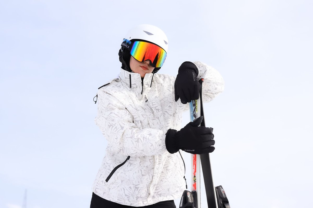 Stylish skier in white winter jacket and mirrored goggles holding skis against a snowy backdrop.