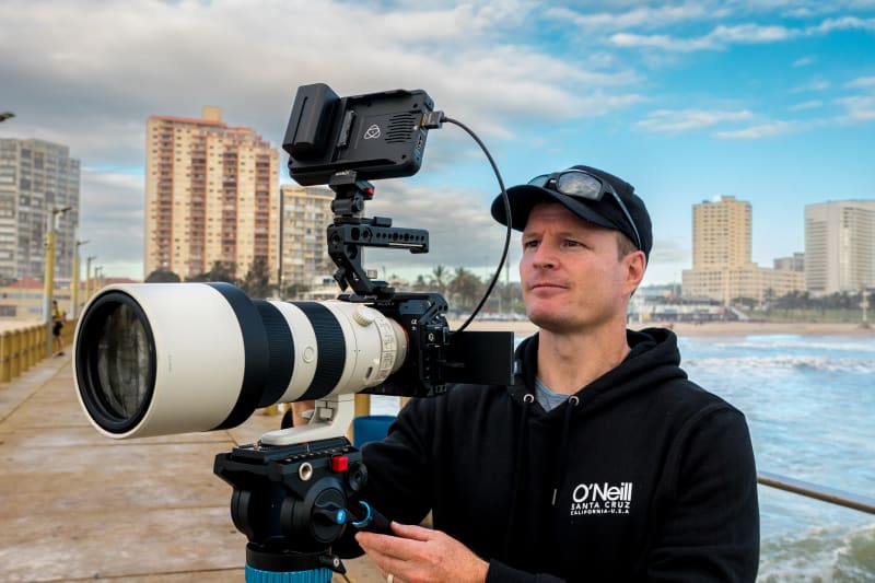 Kevin Rom filming on a Durban pier with a telephoto lens and external monitor, beachfront towers behind