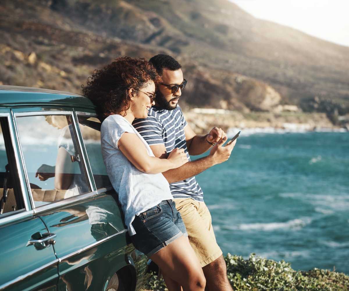 Couple looking at a phone leaning against a car aside the ocean