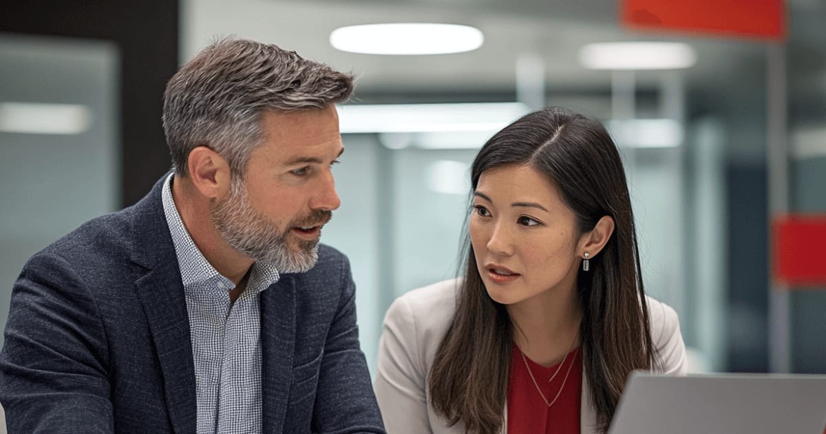 A bearded man in a navy blazer and a woman in a white blazer discussing in front of a laptop in a modern office.