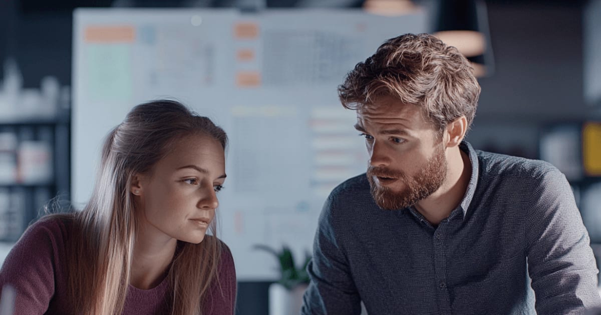 A man and a woman discuss a technical blueprint in an office with a whiteboard in the background.