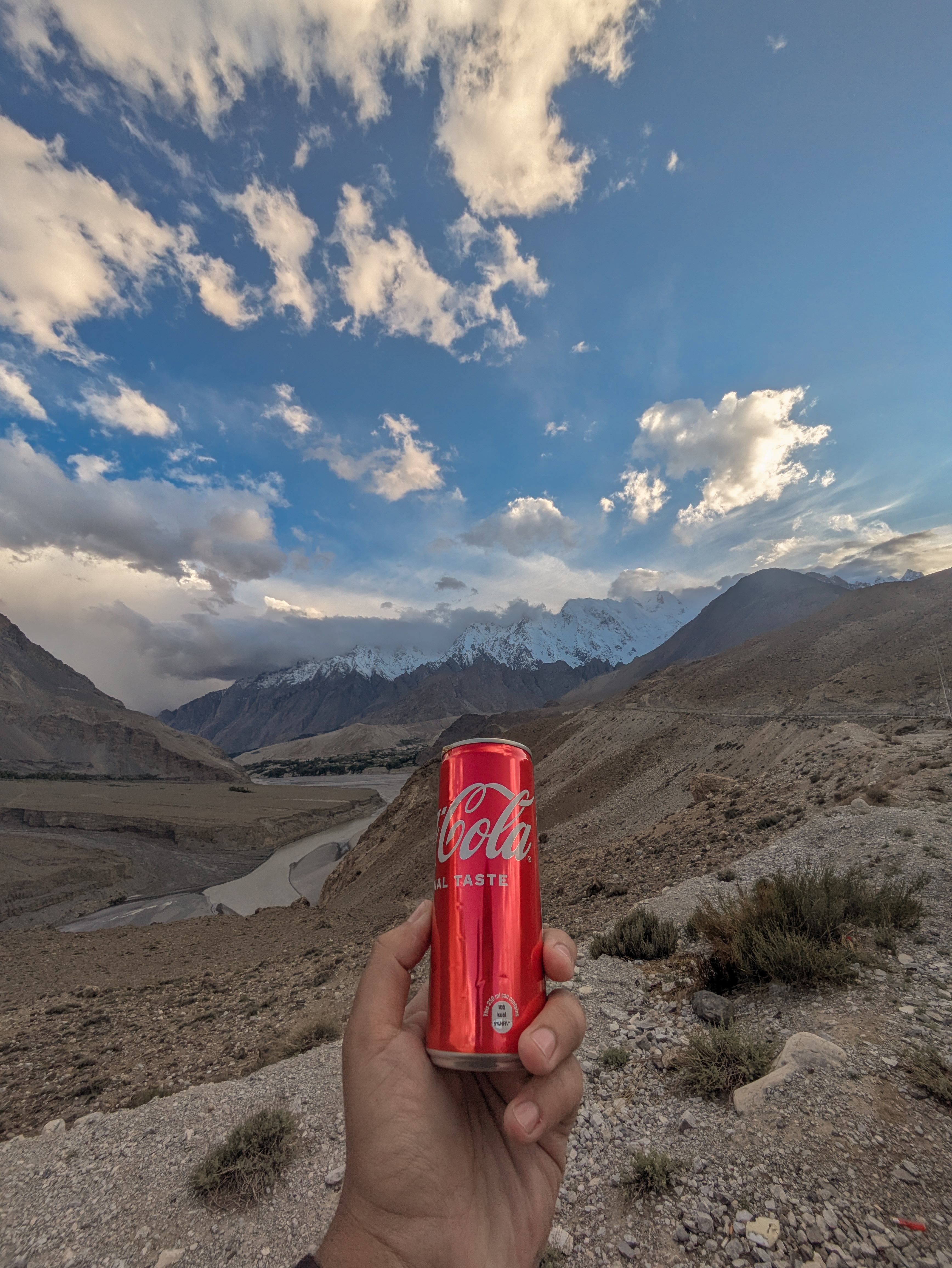 Samie Ullah Baig holding a Coke can with mountains beneath a cloudy sky.