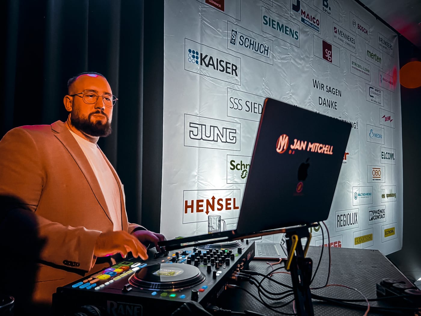 DJ Jan Mitchell am DJ-Pult während der Programmphase einer Firmenfeier im Raum Mannheim mit Sponsoren-Wand im Hintergrund