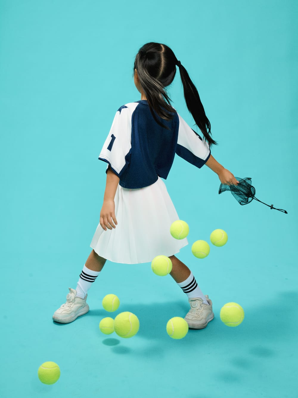 Turned away from camera, swinging an empty ball bag as tennis balls scatter across the floor.