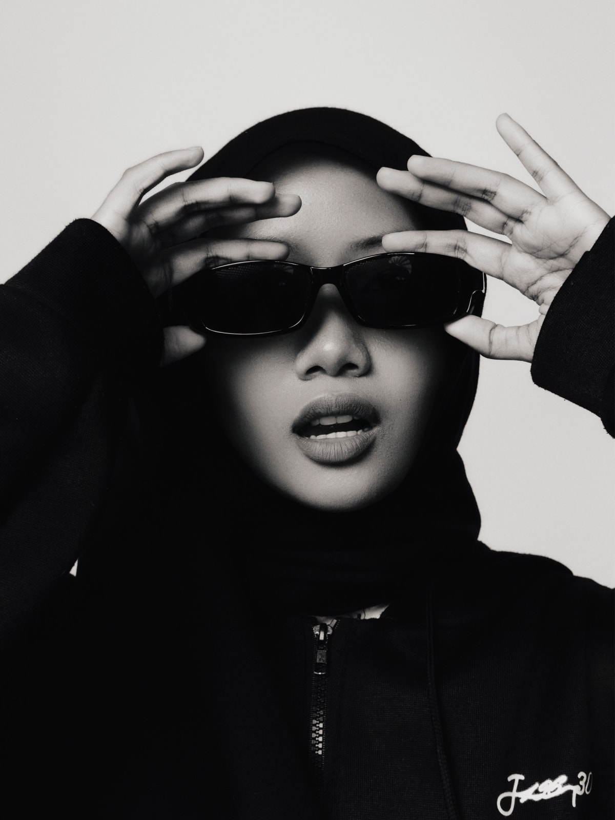 Close frame, both hands raised to frame dark rectangular sunglasses, mouth open, hood up.