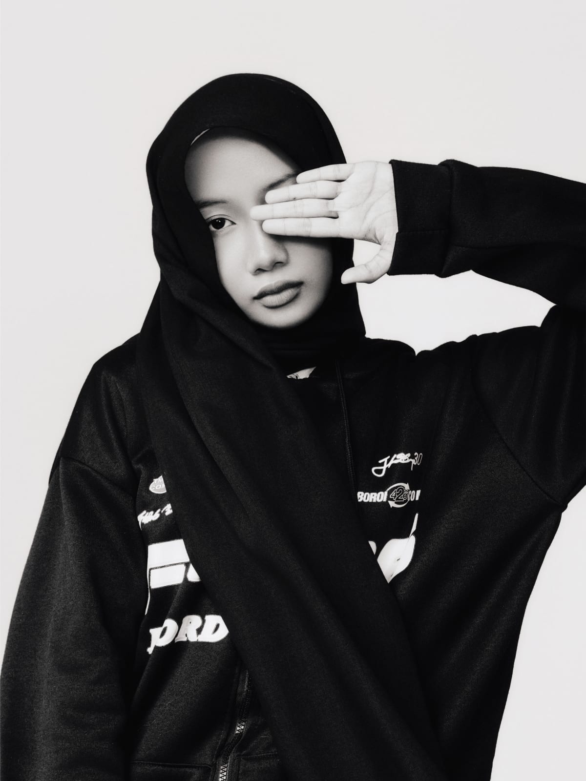 Quiet frame, one hand over an eye, single eye holding the camera, hoodie graphic legible across the chest.