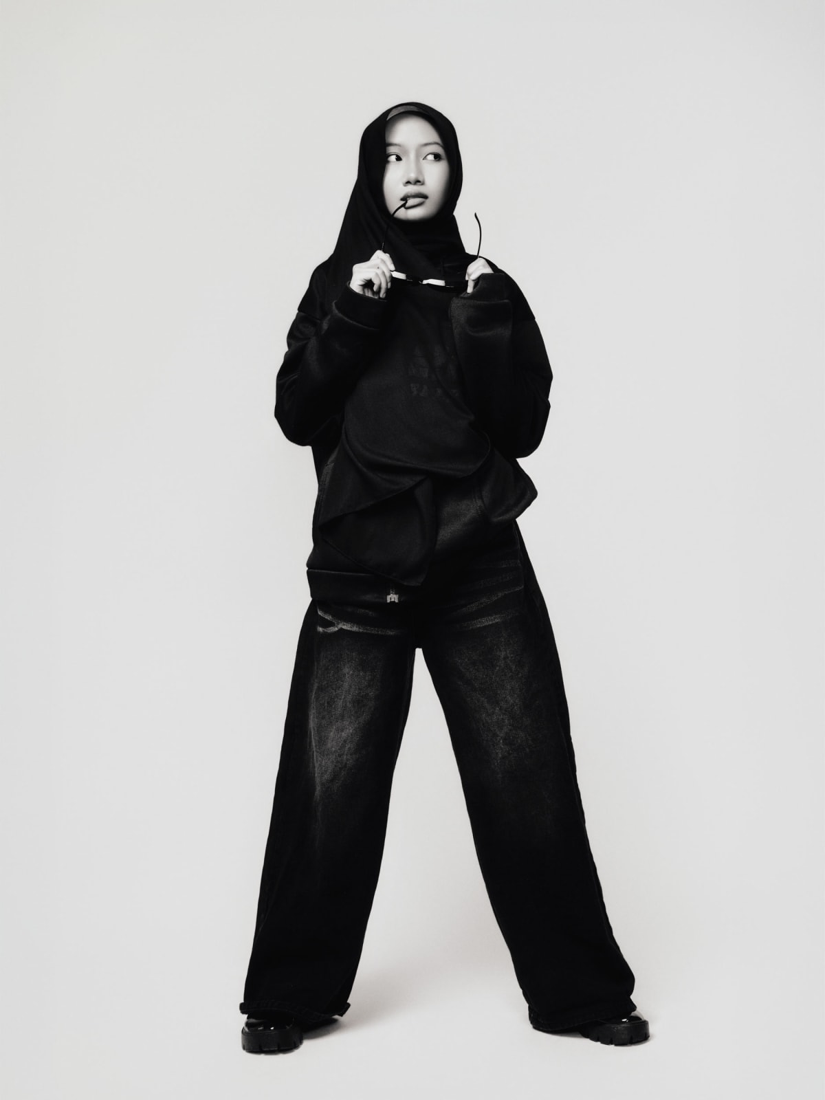 Full-length, standing wide in oversized black hoodie and distressed wide-leg denim, holding sunglasses to her mouth.