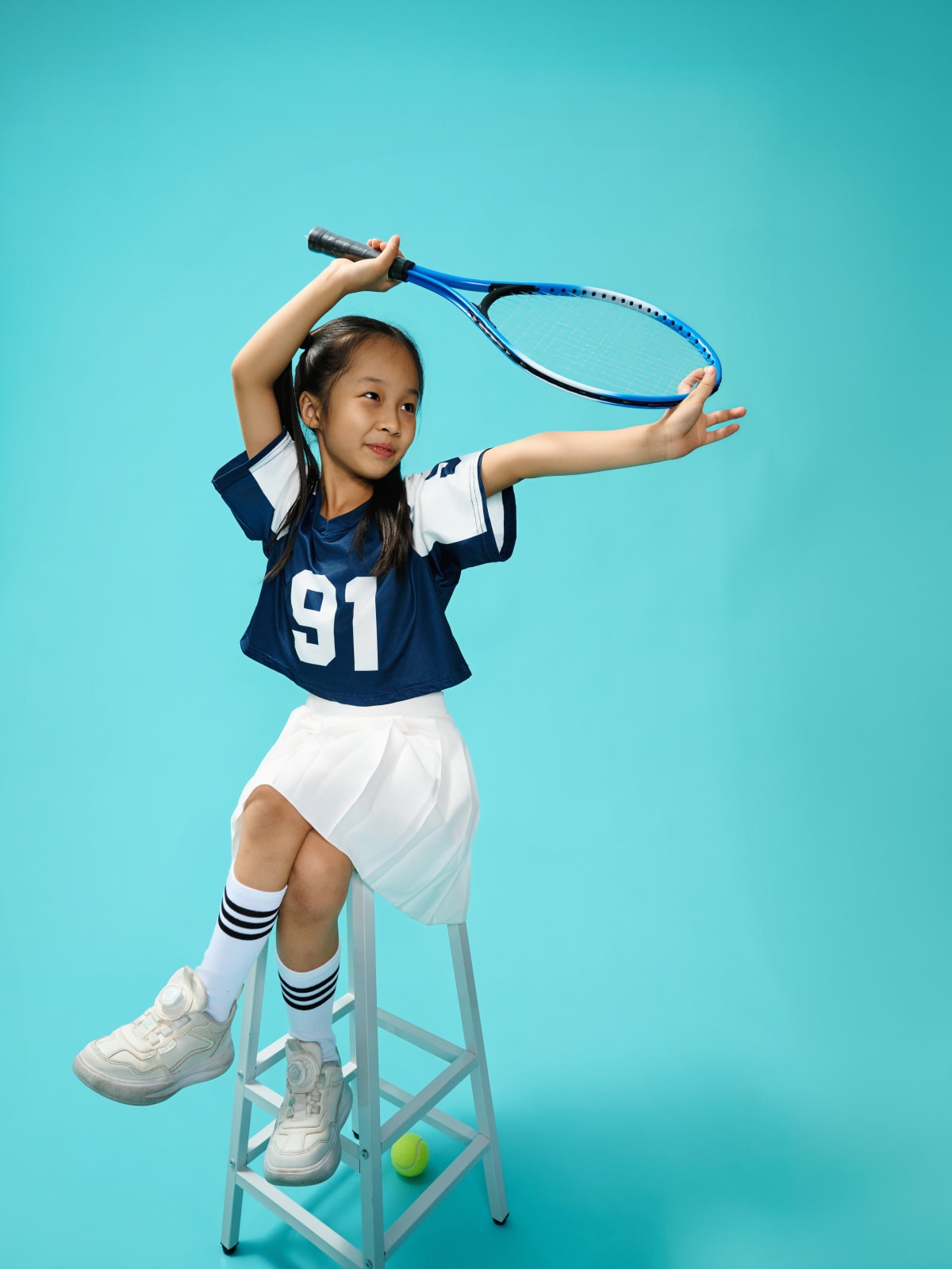 Mid-serve, racket raised overhead, one foot up on a stool against aqua.