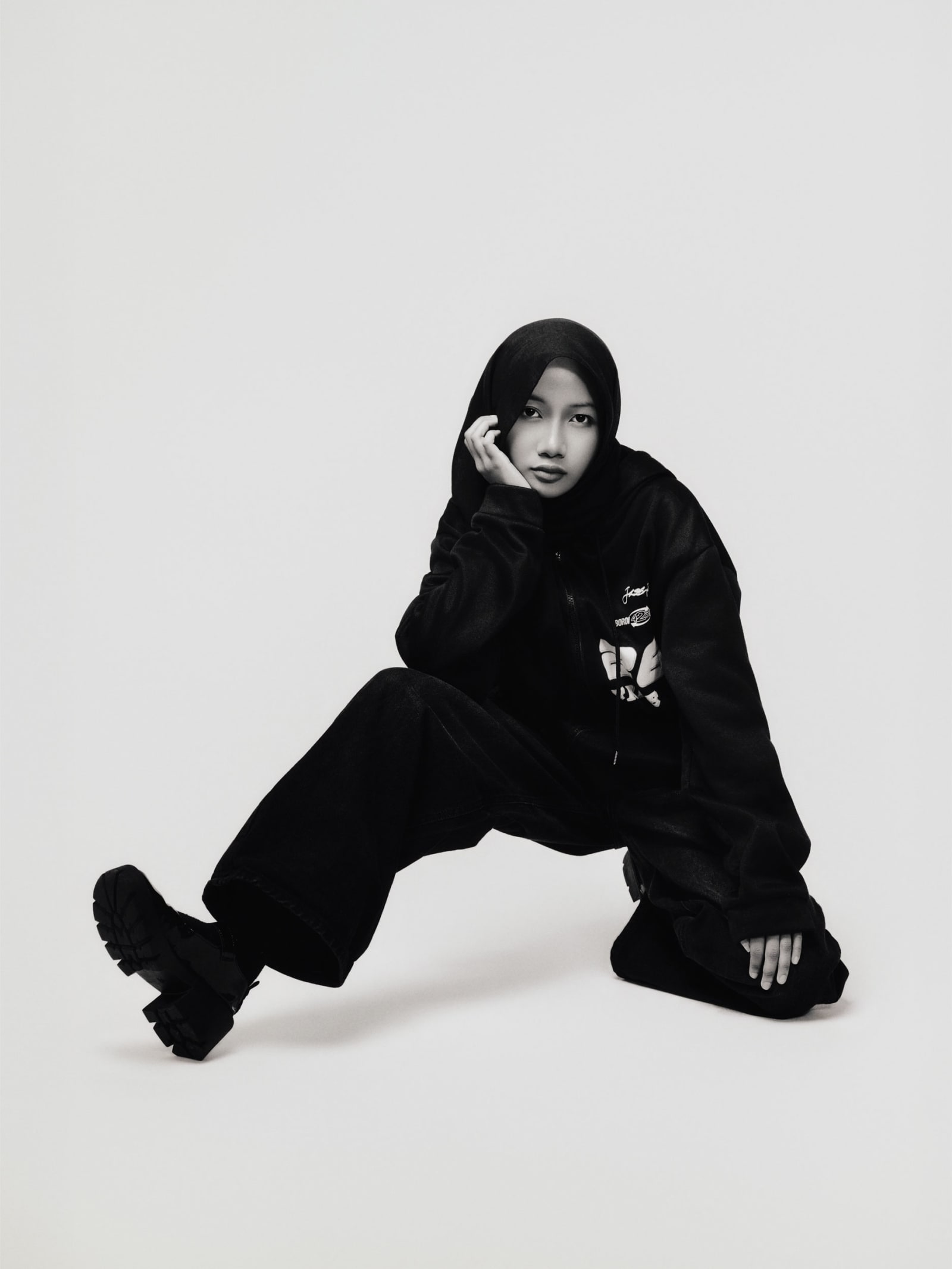Seated on the floor in black hooded streetwear and chunky boots, hand to cheek, holding the camera with a level stare.