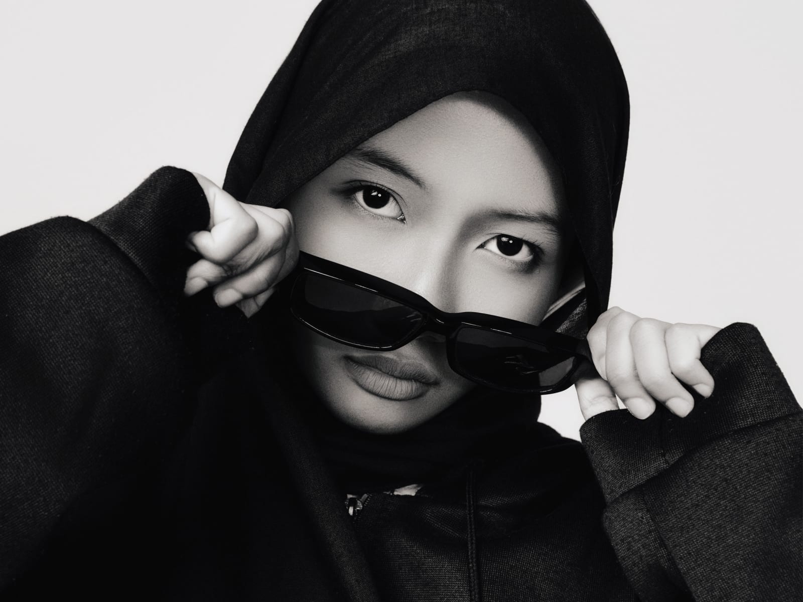 Close crop, hood tugged with both hands, sunglasses low on the nose, eyes holding the camera over the frames.