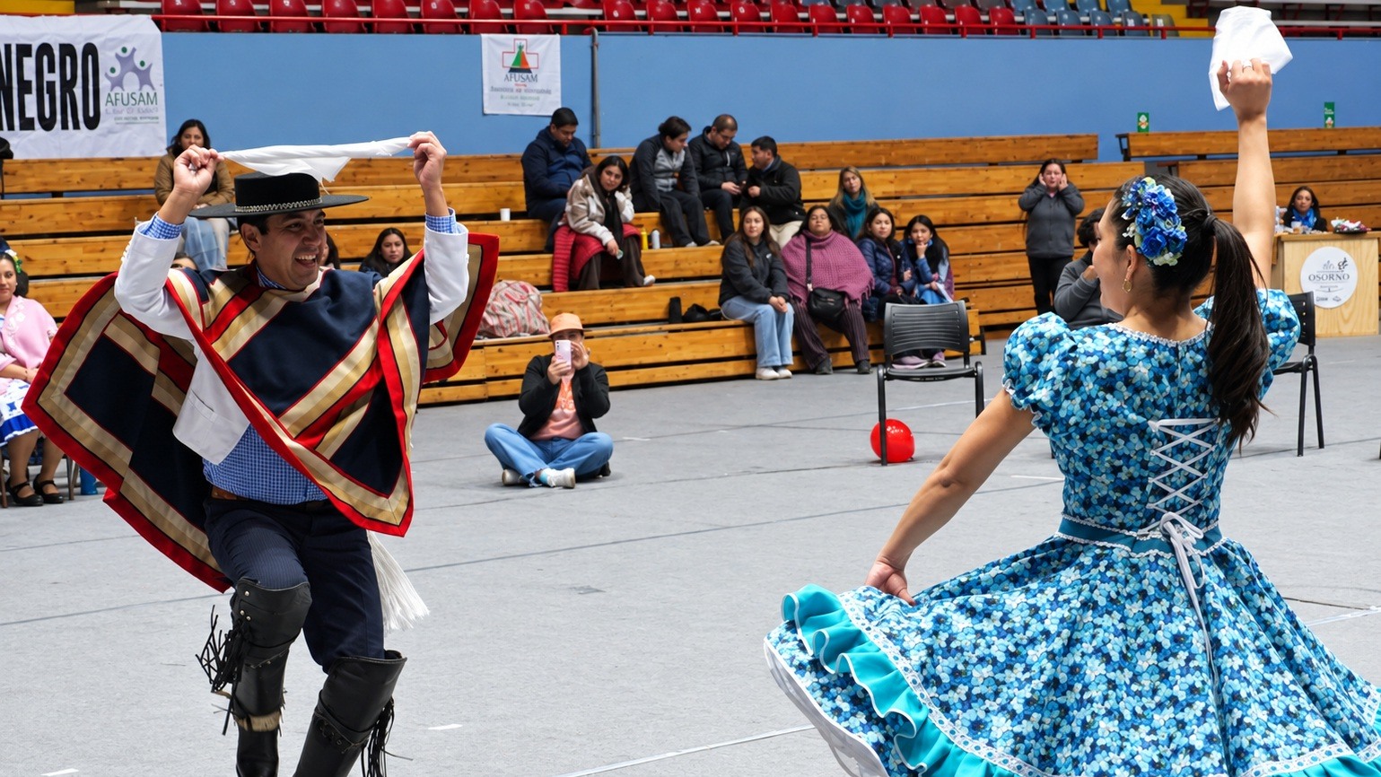 Una pareja baila cueca con panuelos en alto en el gimnasio Maria Gallardo de Osorno, ella con vestido azul floreado y el con manta y sombrero